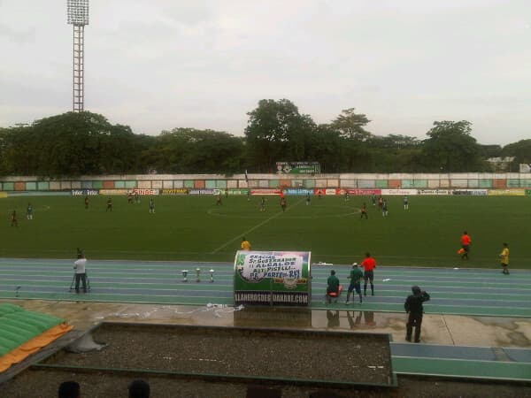 Ein kolumbianisches Fußballstadion vor dem Spiel zwischen Llaneros FC und América de Cali, mit Fans und gepflegtem Rasenplatz.